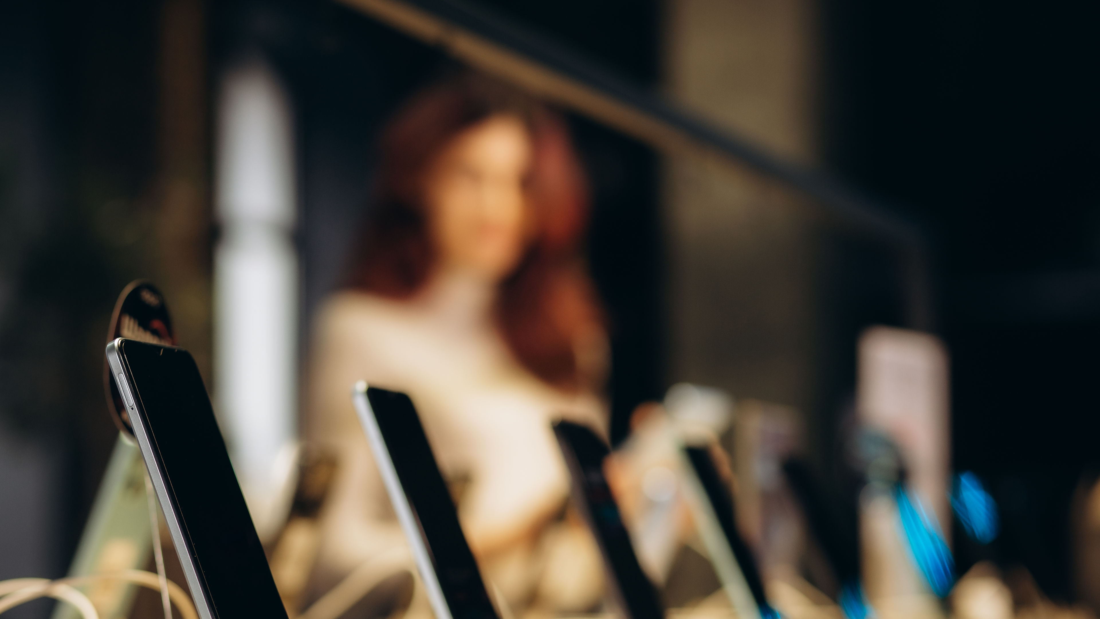 A row of mobile phones mounted on holders in the foreground, with a blurred image of a girl standing in the background.