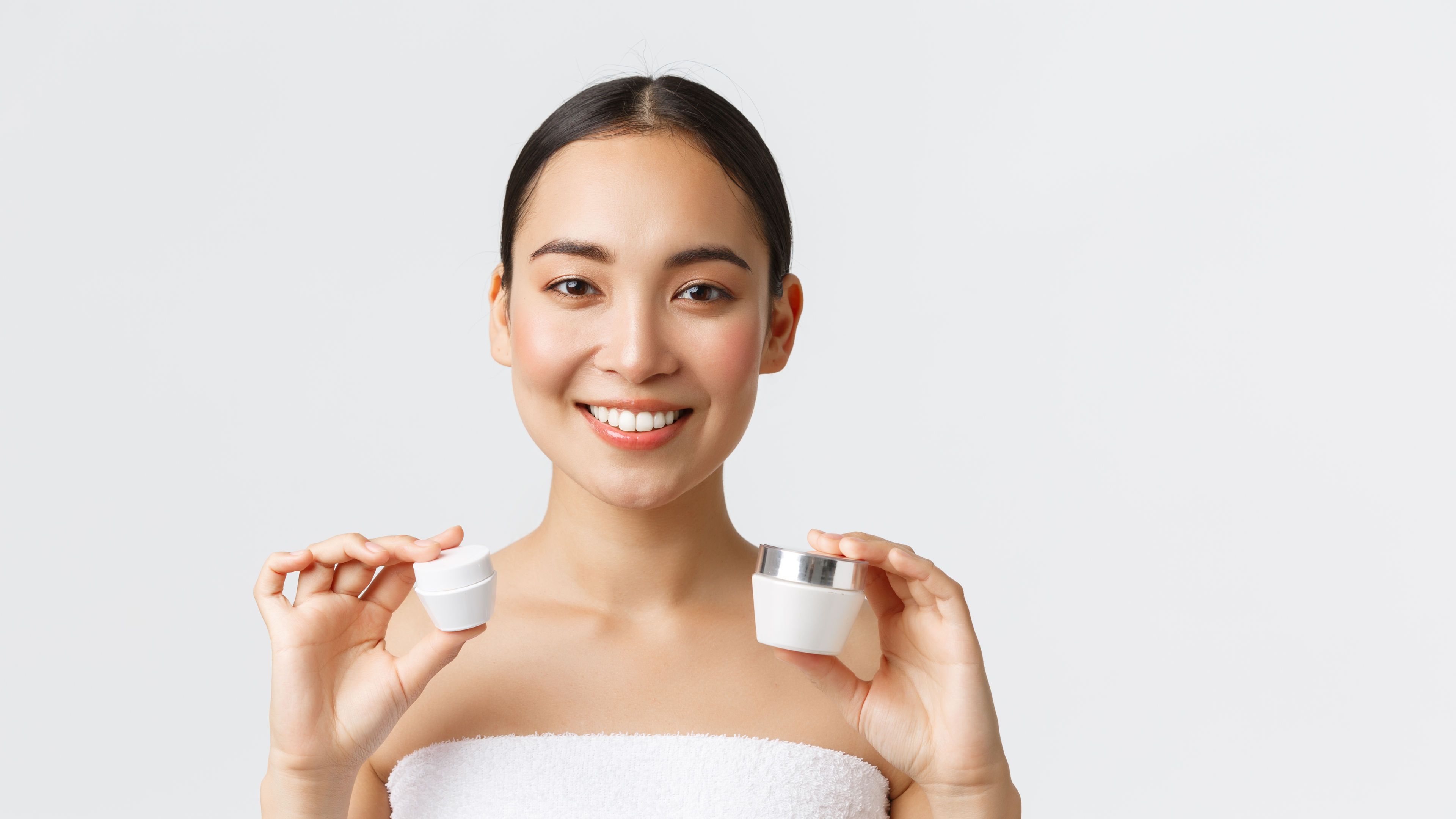 Beautiful Asian woman in bath towel showing facial and eye creams.