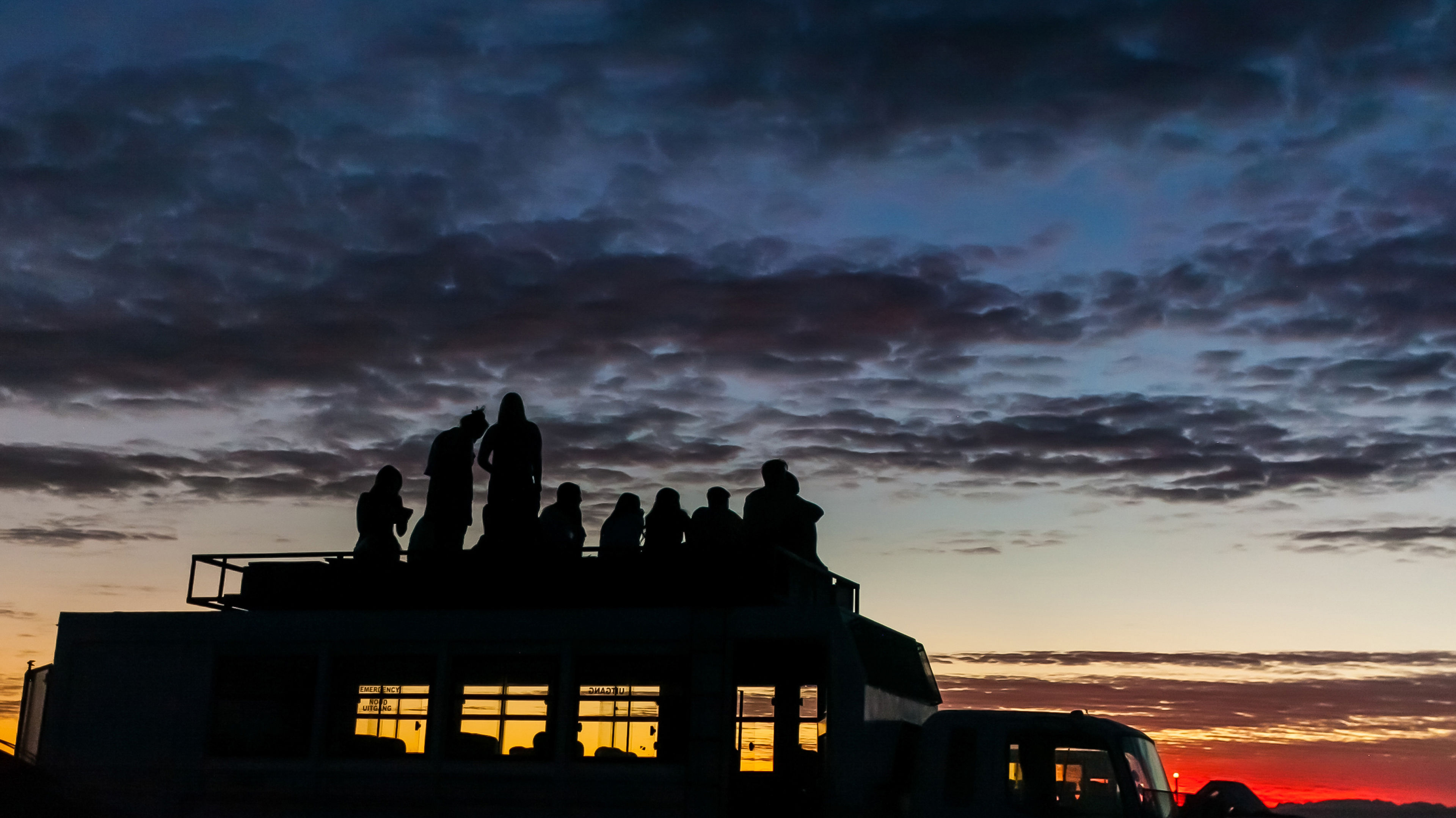A group of people is sitting and standing on the rooftop of a bus