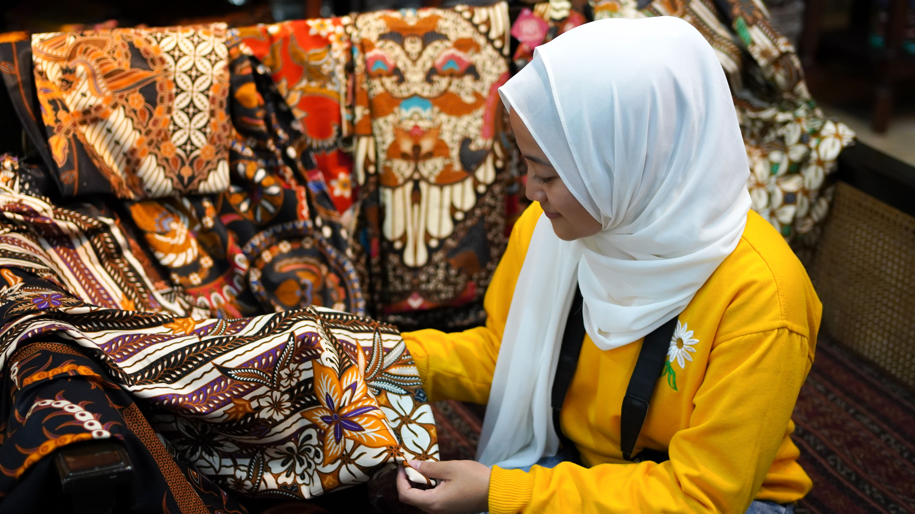 A woman in a white hijab and yellow sweater admires vibrant batik fabric in a marketplace. 