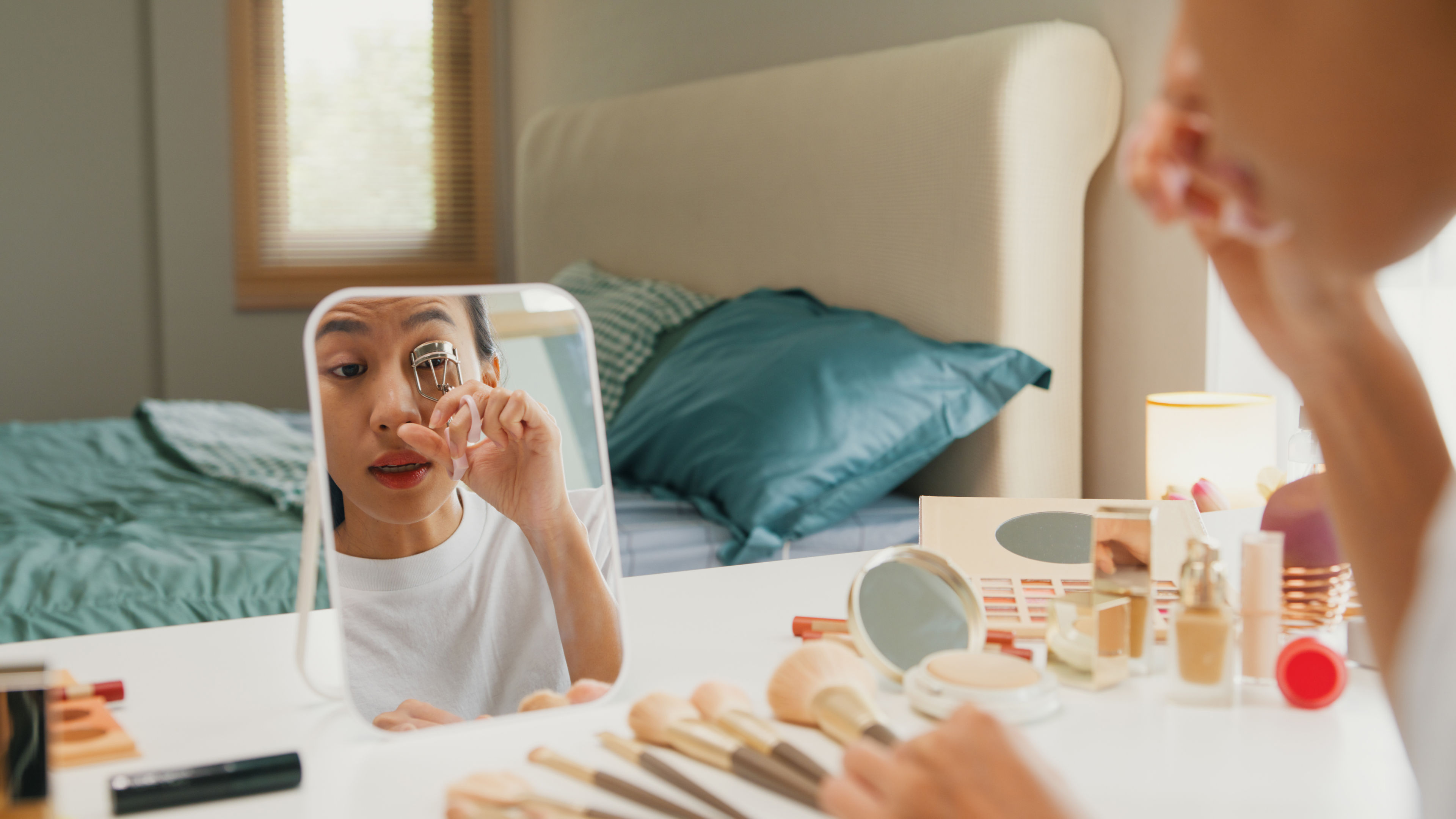 A woman is applying makeup in front of a small mirror with beauty products on the table