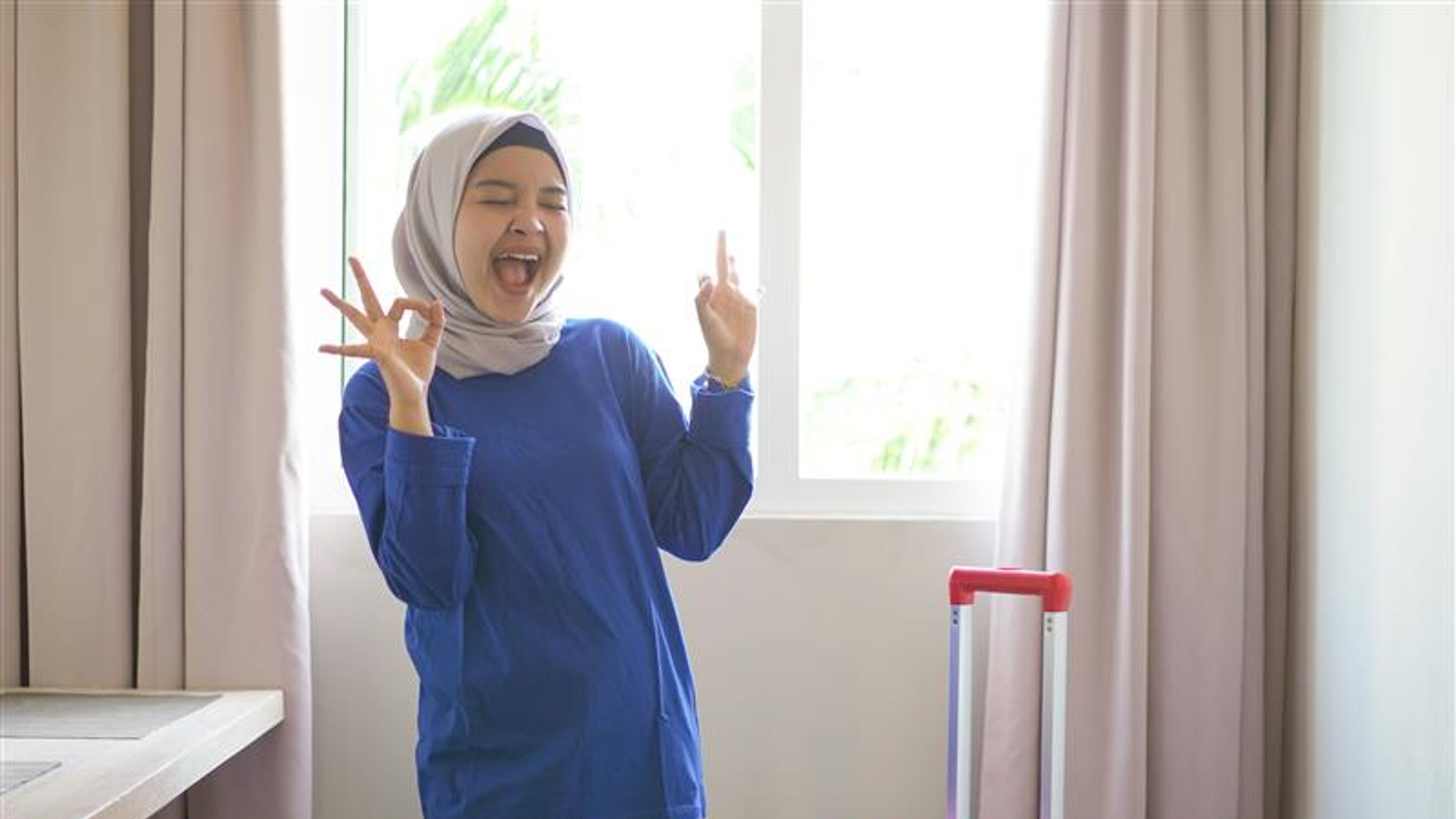 wanita hijab asia ceria memakai kemeja-t biru dan mengangkat tangan dengan isyarat okey di sebuah hotel