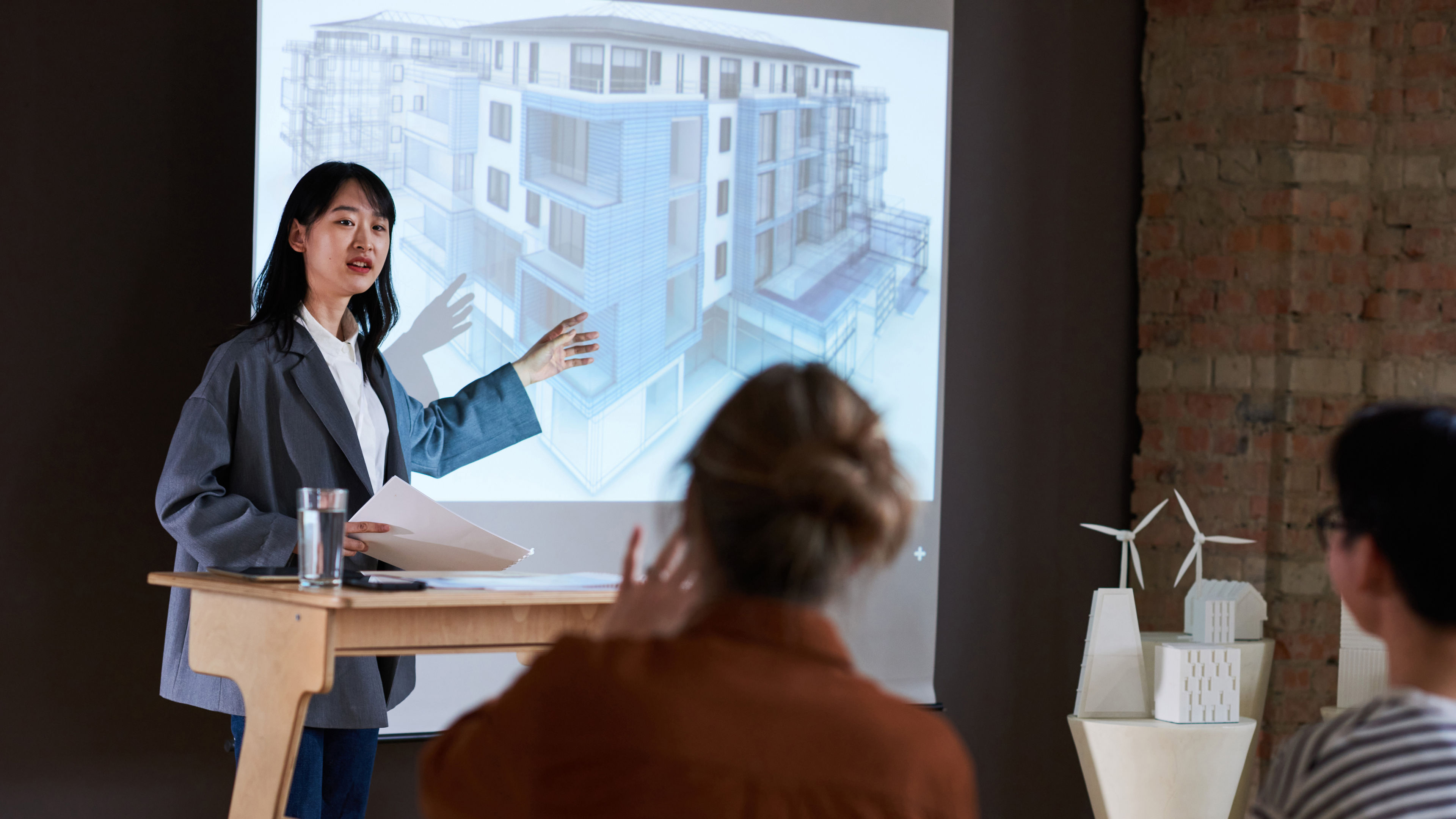Young businesswoman pointing at architectural blueprint and presenting new project to people during a seminar.